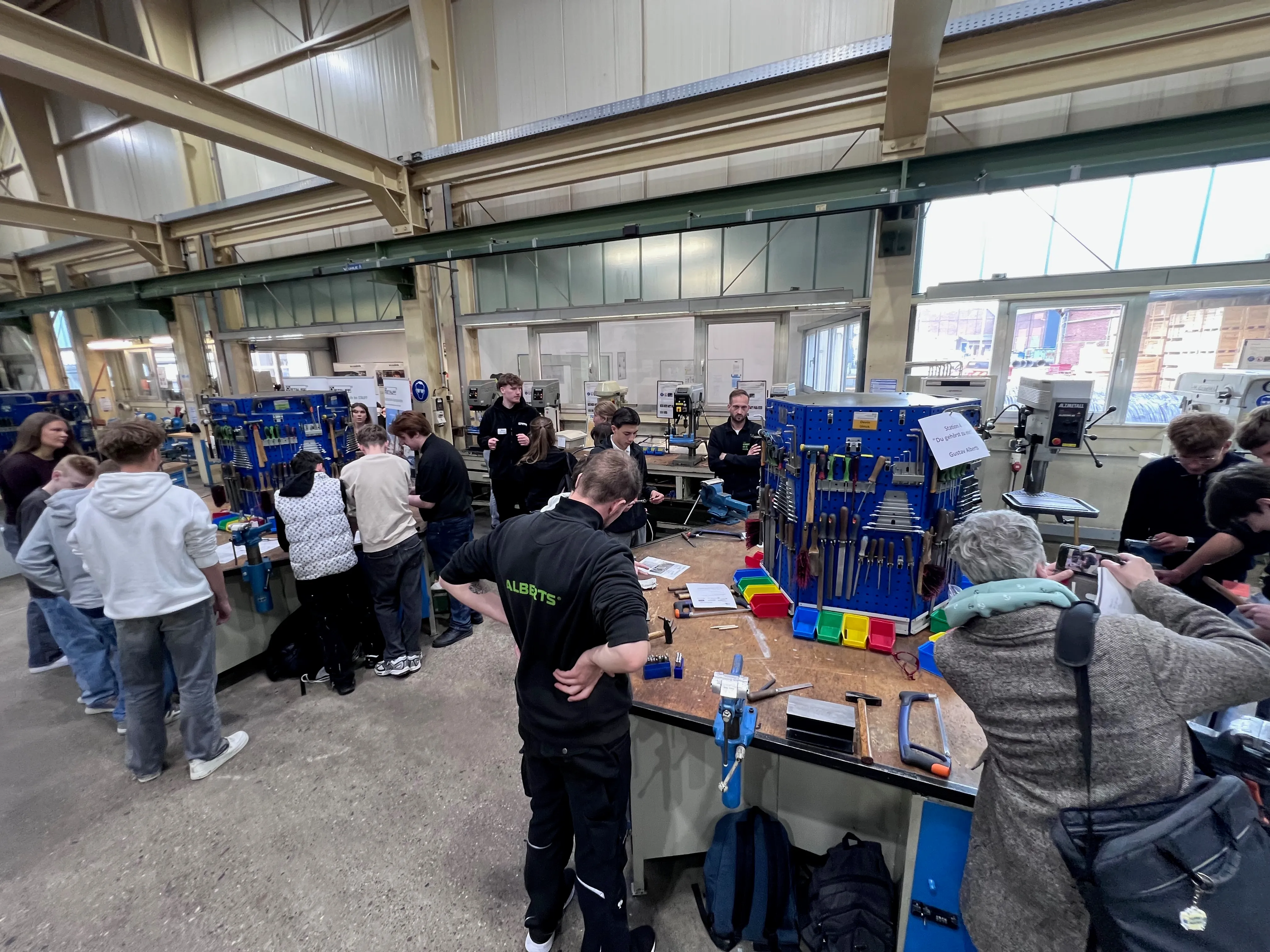 Schülergruppen probieren an verschiedenen Werkstationen handwerkliche Aufgaben aus, darunter das Bearbeiten von Metall und Holz. Im Hintergrund sind weitere Schüler und Auszubildende sowie Werkzeuge und Maschinen zu sehen.