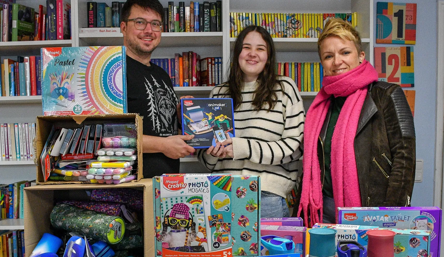 Drei Personen stehen vor Regalen mit Büchern und einer Auswahl an Bastelmaterialien. Die Spenderin Emely Blaß und Cathrin Baumann überreichen dem Leiter der Bildungsstation Benjamin Breuninger eine Spende von Maped Deutschland.