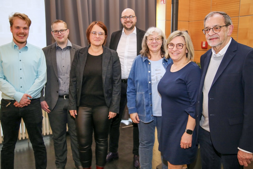 Sieben Personen posieren für ein Gruppenfoto. Von links: ein junger Mann mit Hemd, ein Mann mit Brille und Jacke, eine Frau mit Blazer, ein Mann mit Bart, eine Frau mit Jeanshemd, eine Frau mit blauem Kleid und ein älterer Mann mit Anzug und Brille.