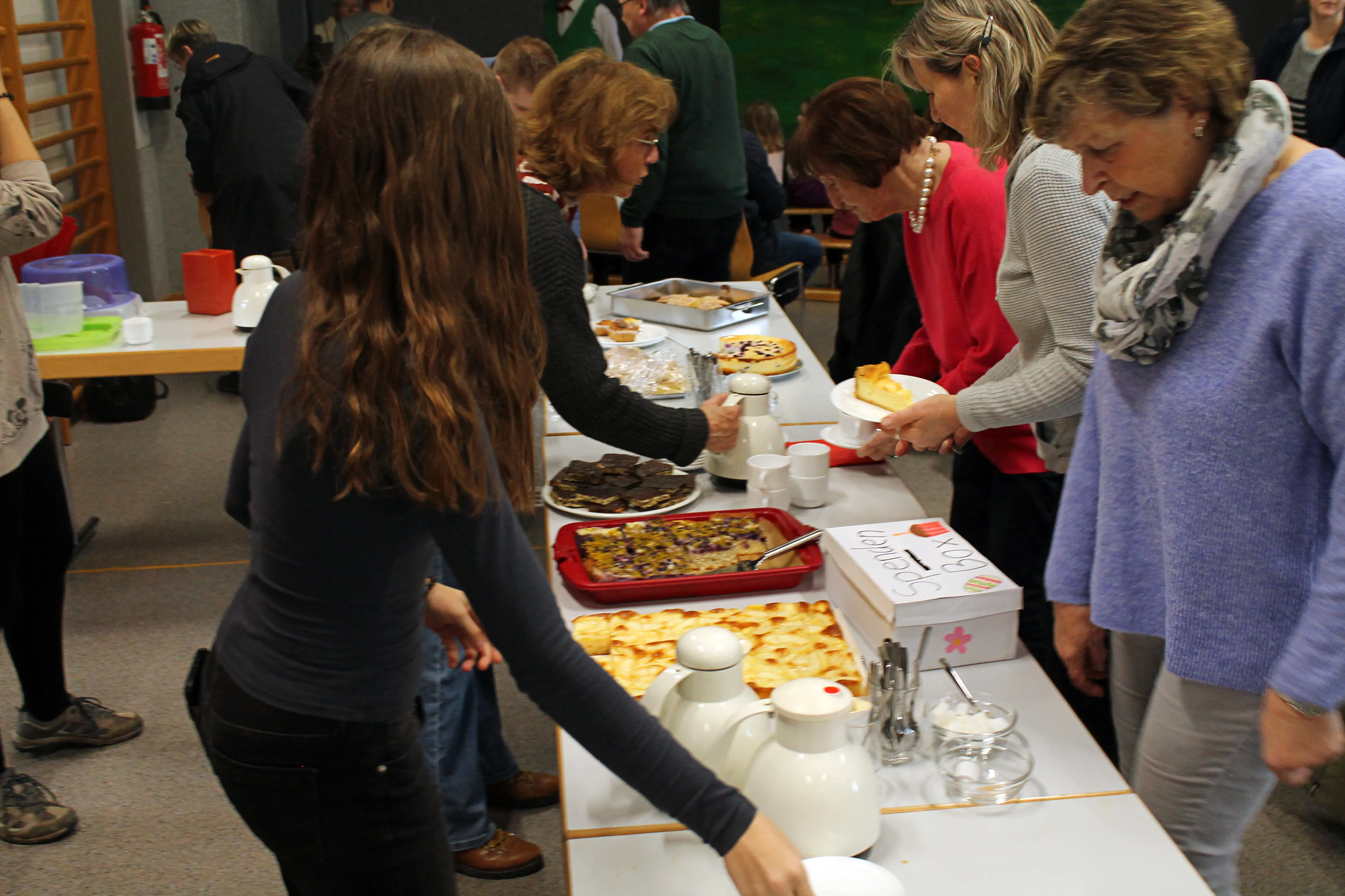 Menschen stehen um einen Tisch mit Speisen, darunter Kuchen und Gebäck, und bedienen sich. Im Hintergrund weitere Personen und ein Blick in den Raum.