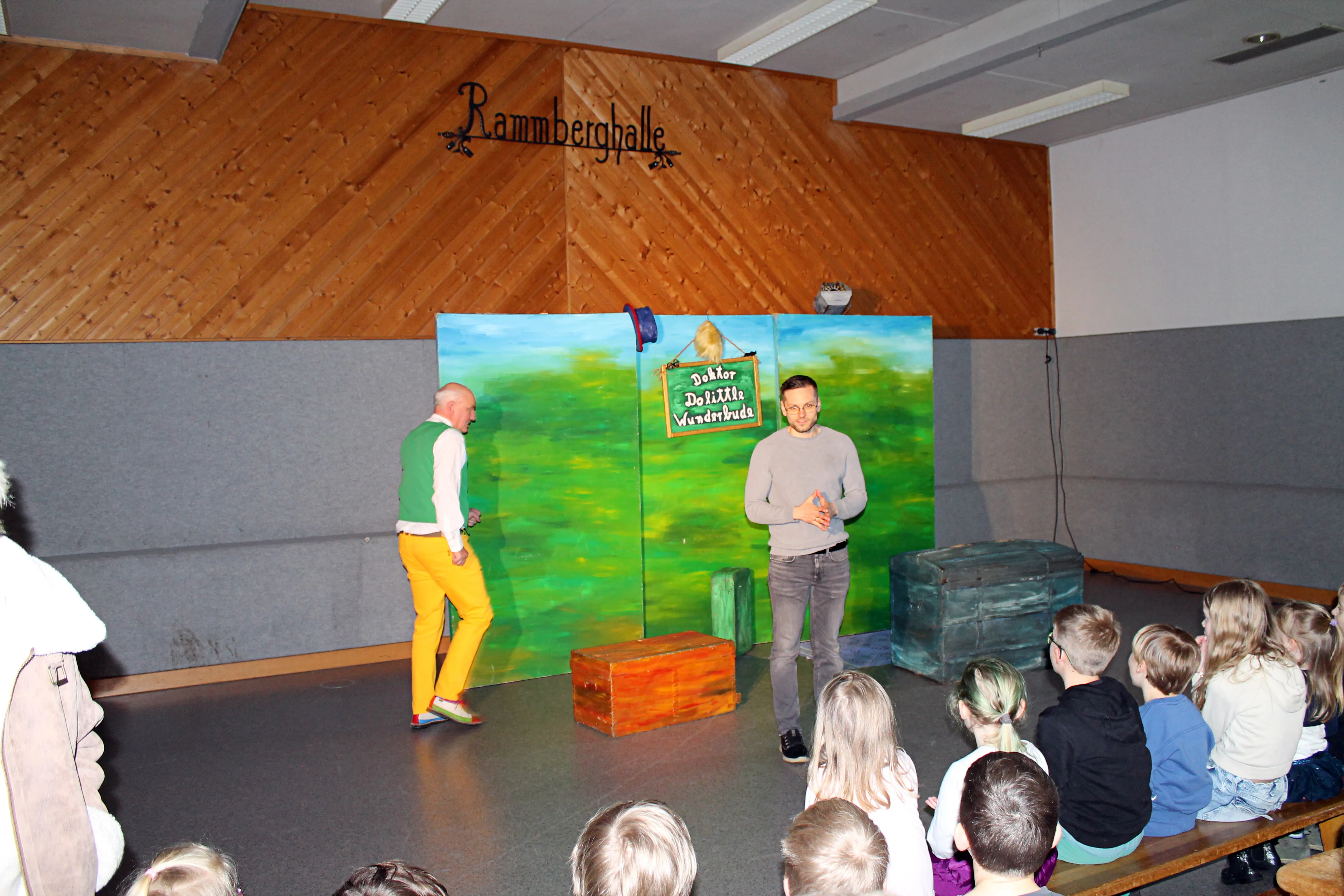 Zwei Männer unterhalten sich vor einer Gruppe von Kindern in einer Holzhalle. Im Hintergrund ein gemaltes Landschaftsbild mit dem Schriftzug „Doktor Drollbarda Wunderbuda“.