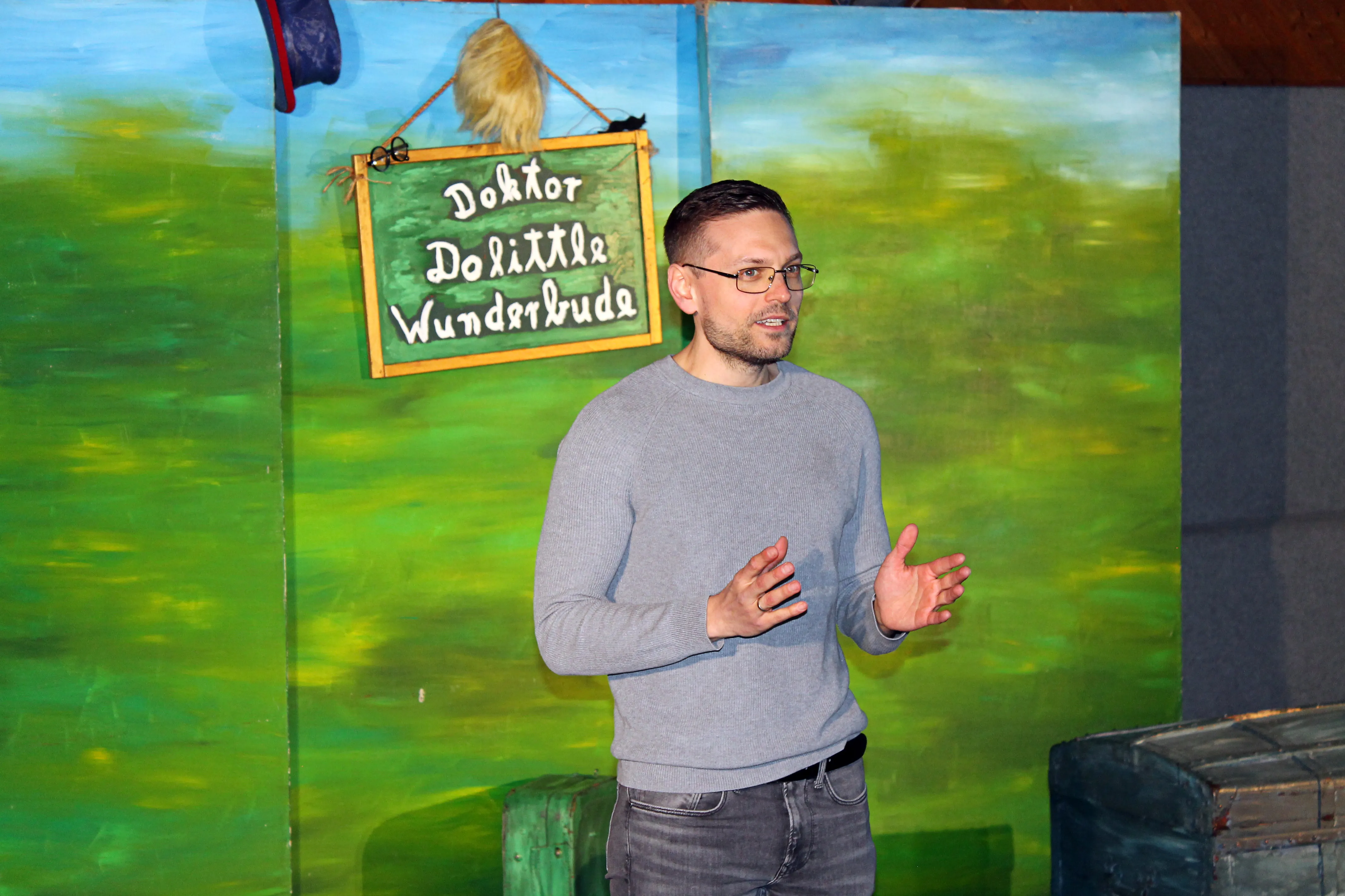 Ein Mann mit Brille und grauem Pullover gestikuliert vor einem Hintergrund mit dem Schild „Doktor Dolittle Wunderbude“ und einem Vogel.