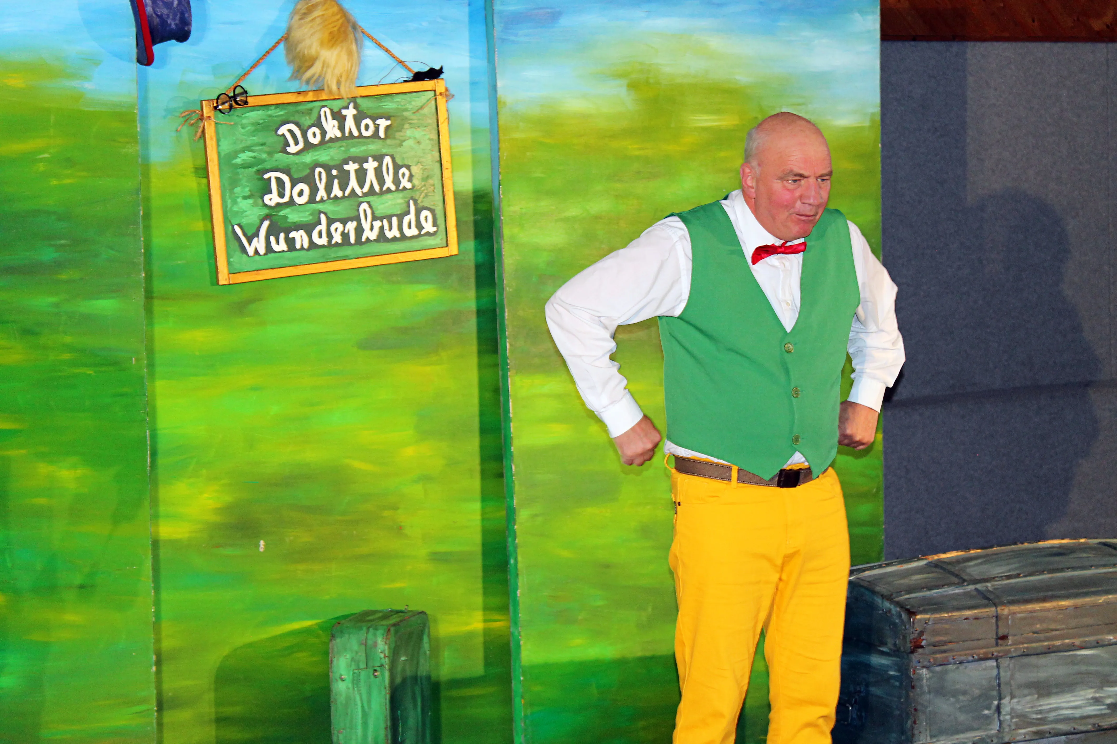 Ein Mann mit Glatze, gelber Hose und grüner Weste steht vor einem Bühnenbild mit dem Schriftzug „Doktor Dolittle Wunderbude“ und hält die Hände in die Hüften.