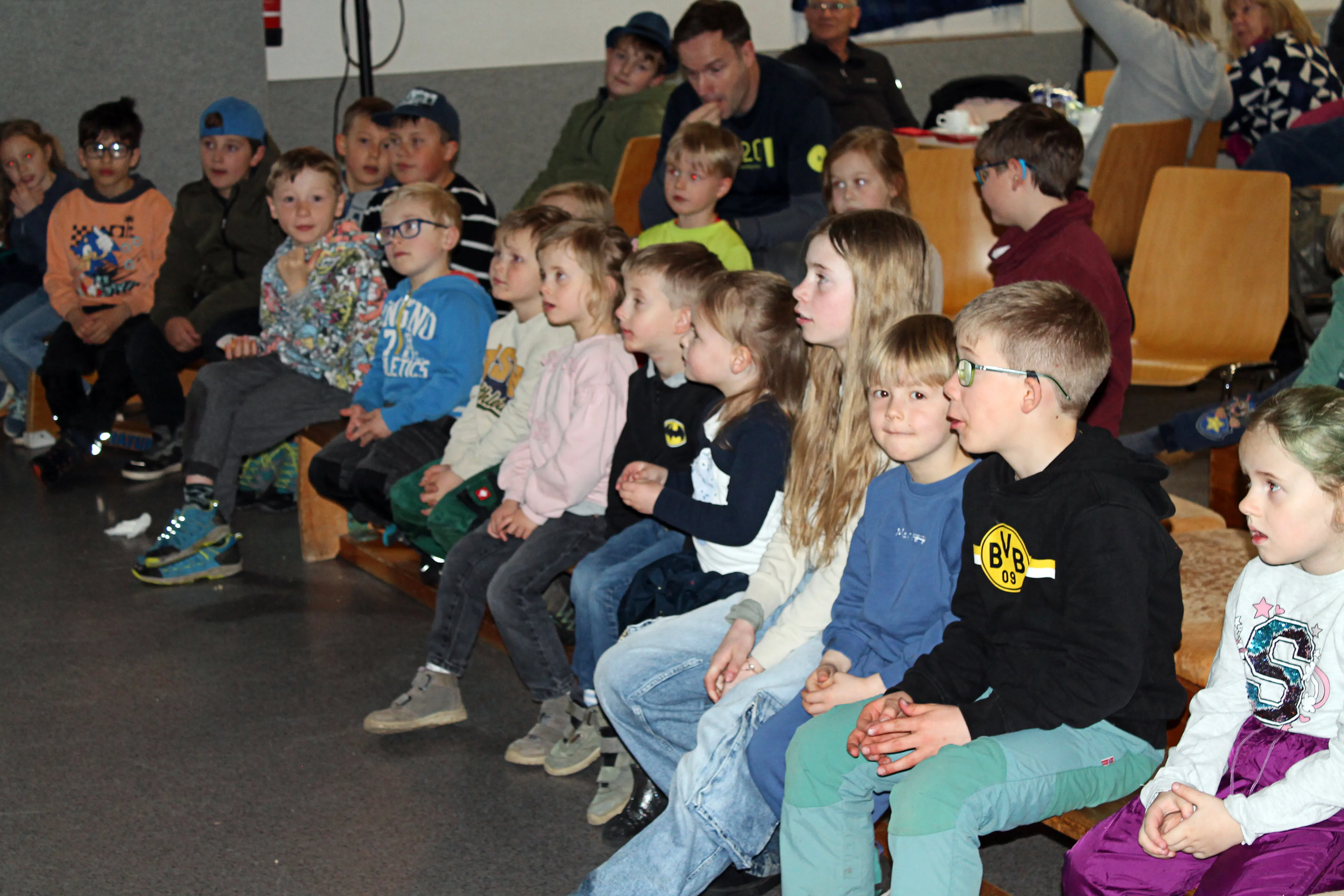 Zuschauer, hauptsächlich Kinder, sitzen auf Bänken und schauen aufmerksam nach vorne, einige tragen farbige Jacken und Pullover.