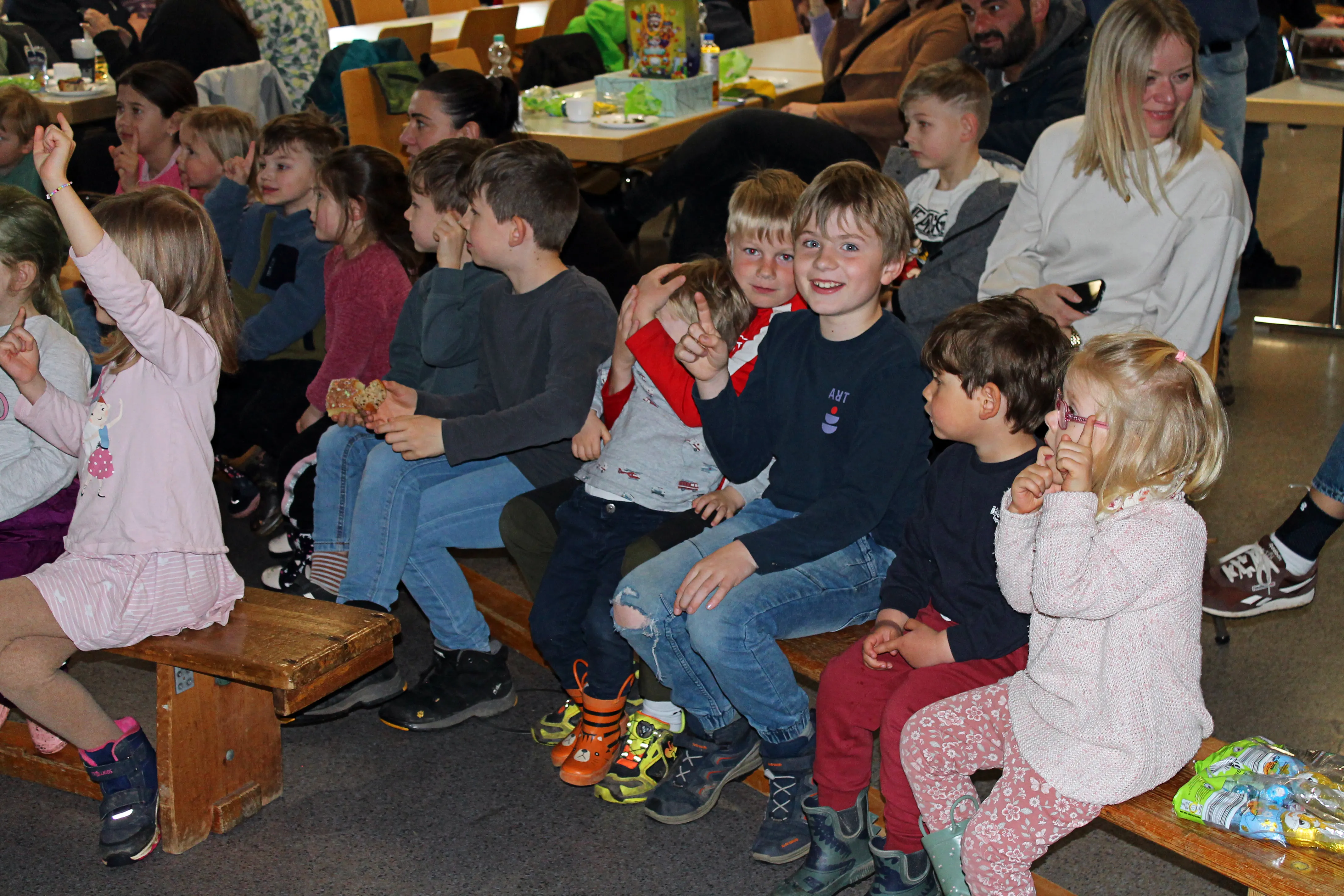 Mehrere Kinder und Eltern sitzen auf Holzbänken und schauen erwartungsvoll nach vorne, einige Kinder halten Ostereier oder Snacks in den Händen.