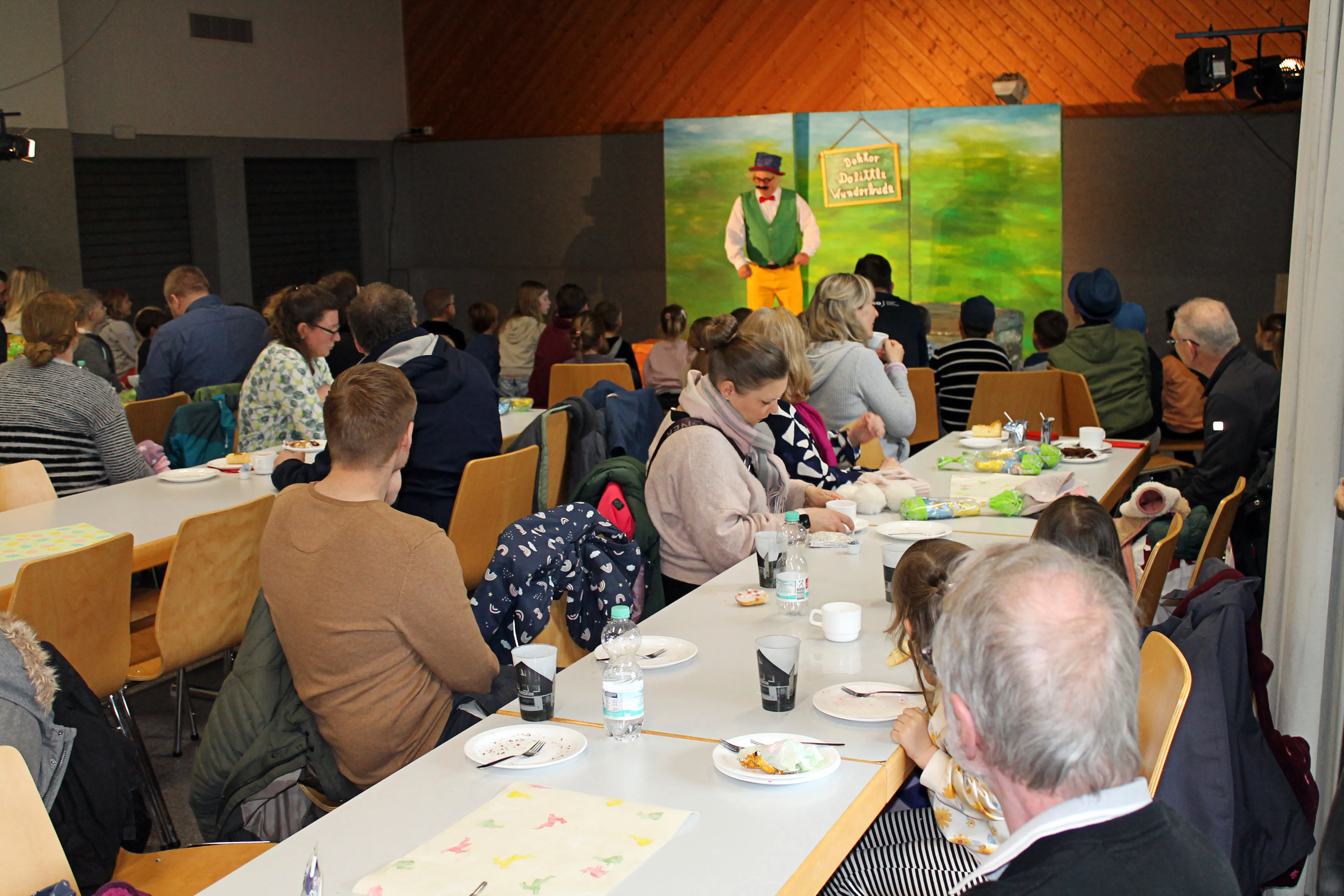 Menschen sitzen an langen Tischen in einer Halle und essen. Im Hintergrund steht ein Mann in Bühnenkleidung vor einer Kulisse mit einem Osterhasen-Motiv.