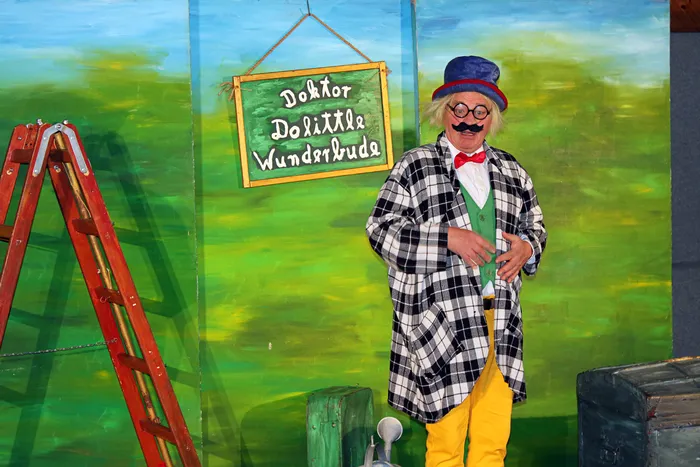 Darsteller mit Schnurrbart und roter Schleife im karierten Jackett, Hut und Brille hält eine grüne Tafel mit der Aufschrift „Doktor Dolittle Wunderbude“.