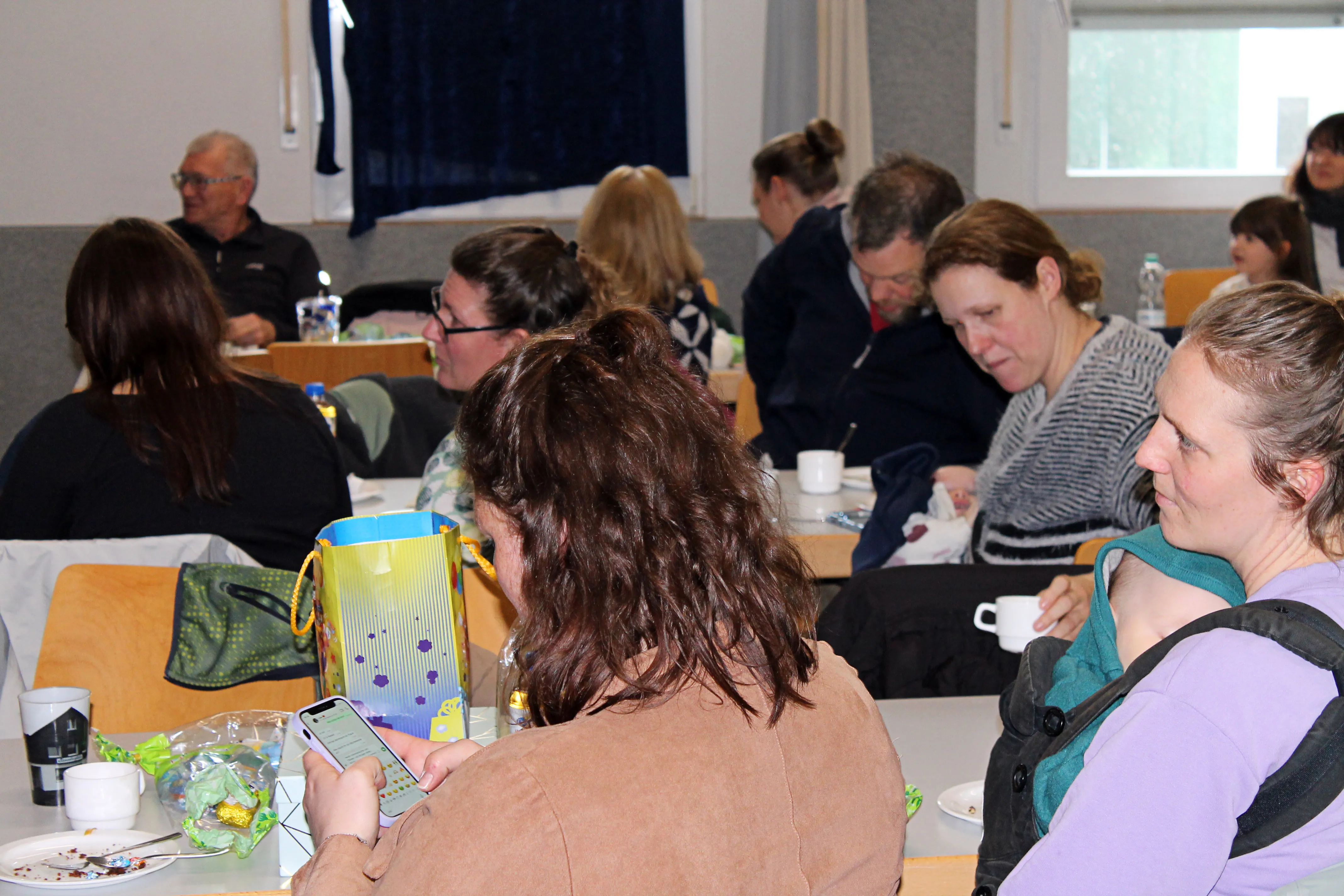 Menschen sitzen an Tischen in einer hellen Halle, einige mit Tassen und Snacks. Eine Frau schaut auf ihr Smartphone, während eine andere ein Baby im Tragetuch hält.
