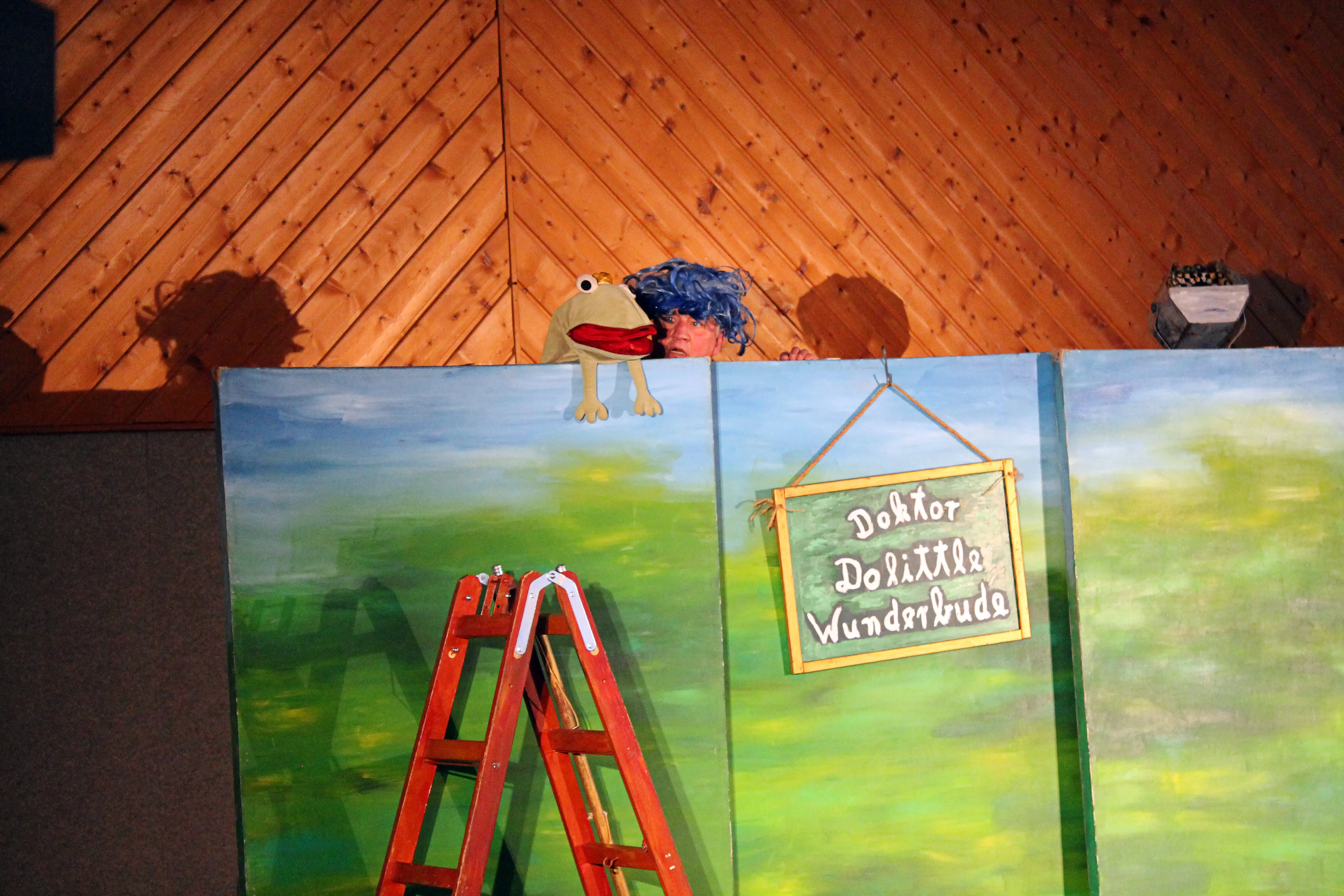 Bühnenbild mit einer grünen Froschpuppe und einem Schild mit der Aufschrift „Doktor Dolittle Wunderbude“. Eine Person mit lockigem Haar ist teilweise hinter dem Bühnenbild zu sehen, eine rote Leiter steht davor.