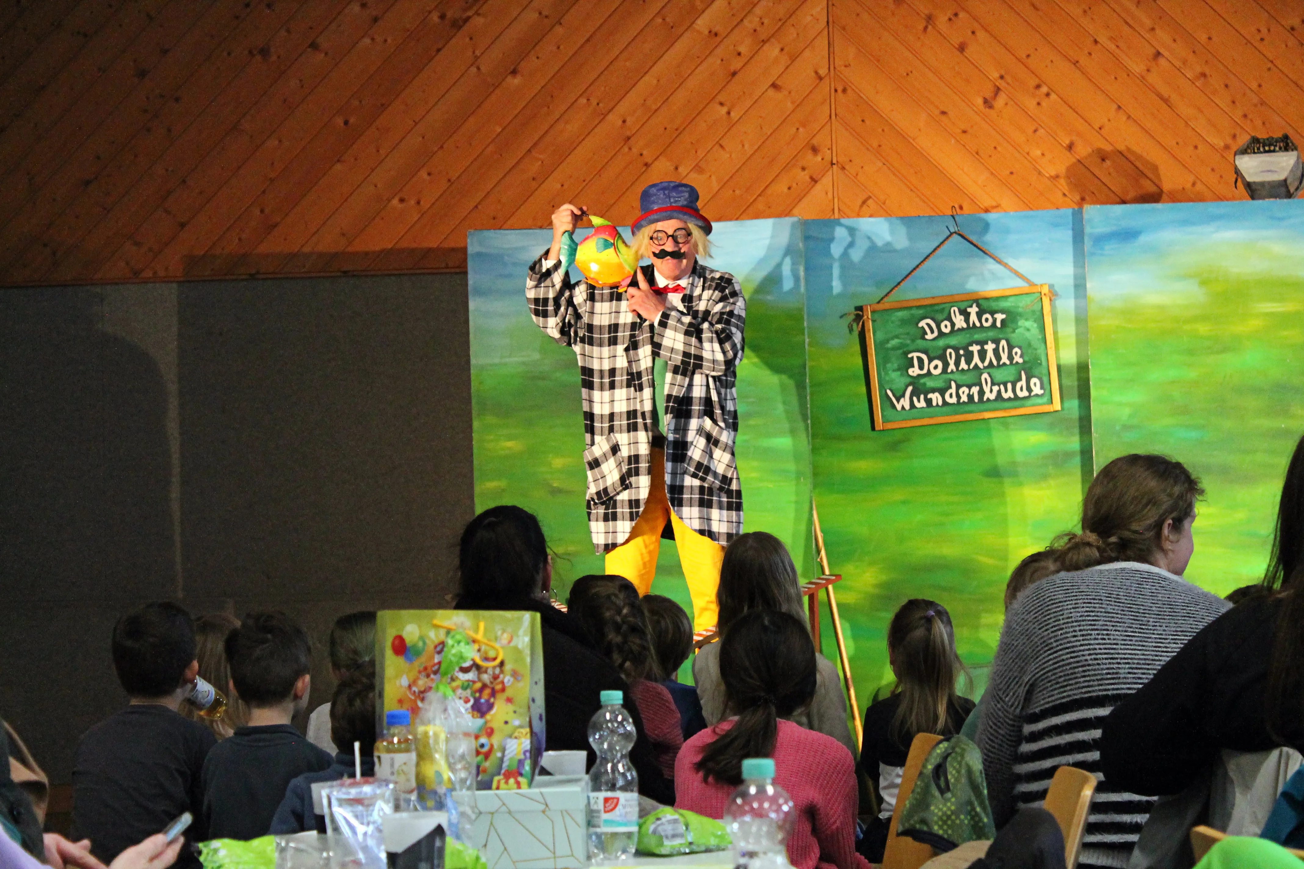 Ein Mann in Verkleidung als Doktor Dolittle mit einem gelben Vogel auf der Hand steht auf einer Bühne vor einem Publikum aus Kindern und Eltern. Im Hintergrund ein grünes Landschaftsbild und ein Schild mit der Aufschrift „Doktor Dolittle Wunderbude“.