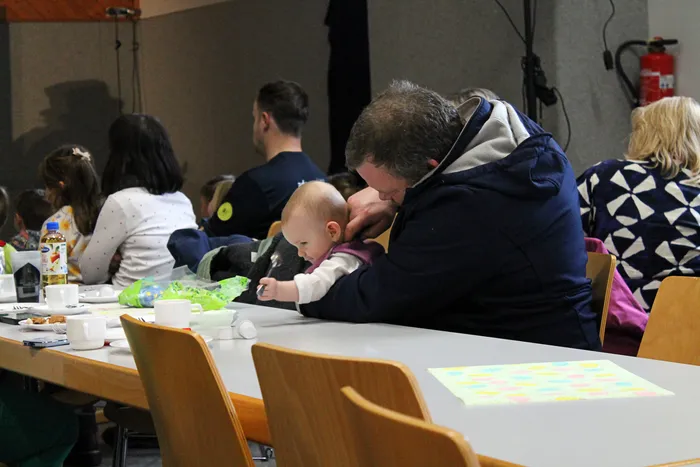 Ein Mann trägt ein Baby auf der Schulter, während sie an einem Tisch mit anderen Familien sitzen. Im Hintergrund sind weitere Personen und ein gedeckter Tisch mit Speisen zu sehen.
