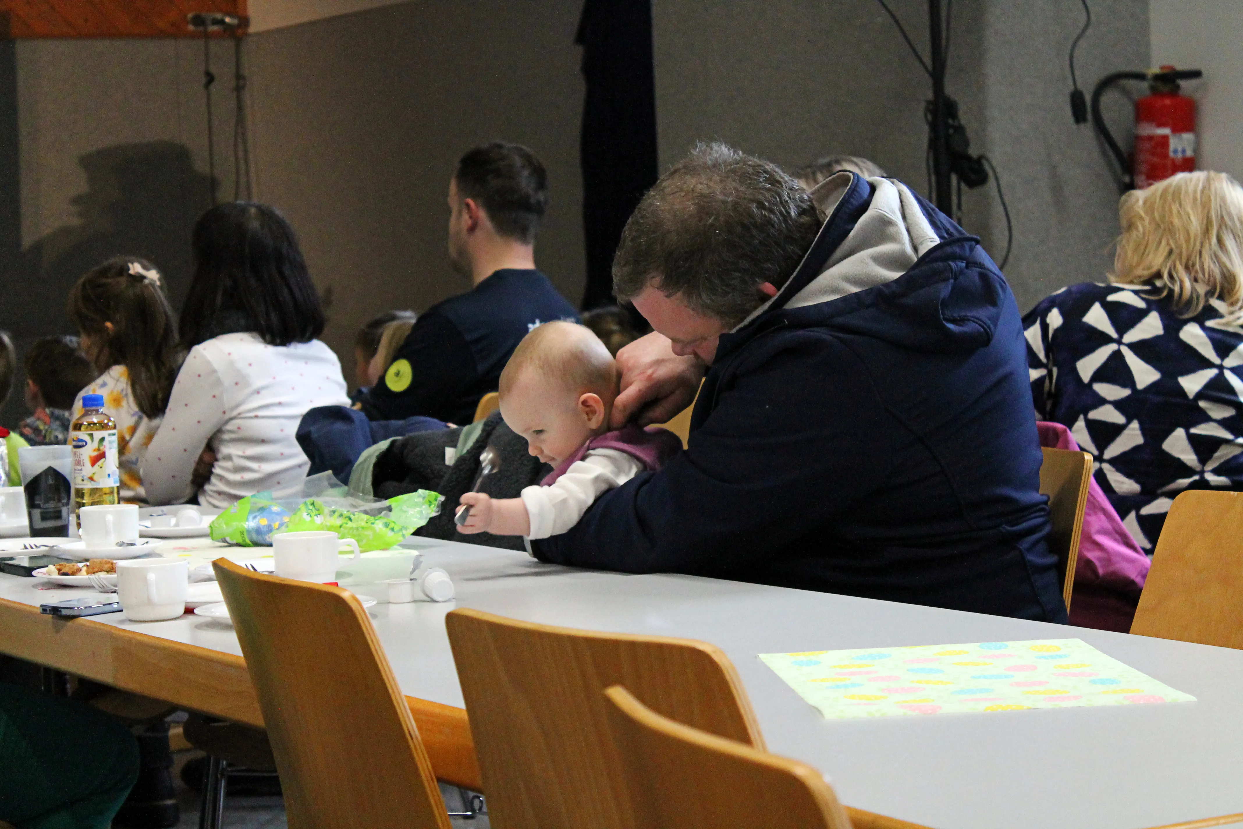 Ein Mann trägt ein Baby auf der Schulter, während sie an einem Tisch mit anderen Familien sitzen. Im Hintergrund sind weitere Personen und ein gedeckter Tisch mit Speisen zu sehen.