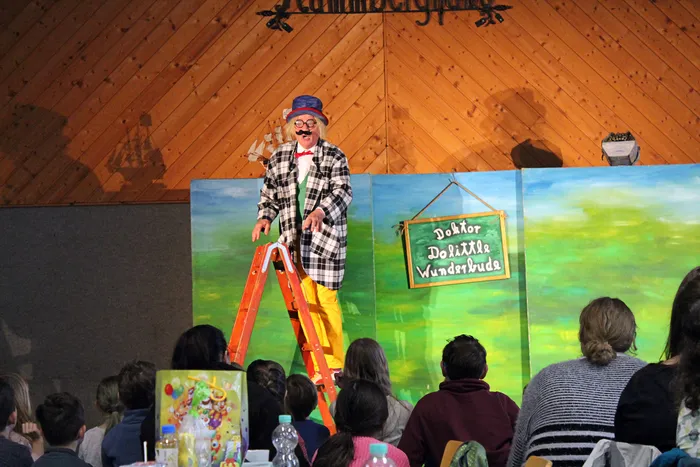 Ein Mann in kariertem Jackett und gelben Hosen steht auf einer Leiter vor einer Kulisse mit Wiesenlandschaft und einem Schild mit der Aufschrift „Doktor Dolittle Wunderbude“. Im Publikum sitzen Kinder und Erwachsene.