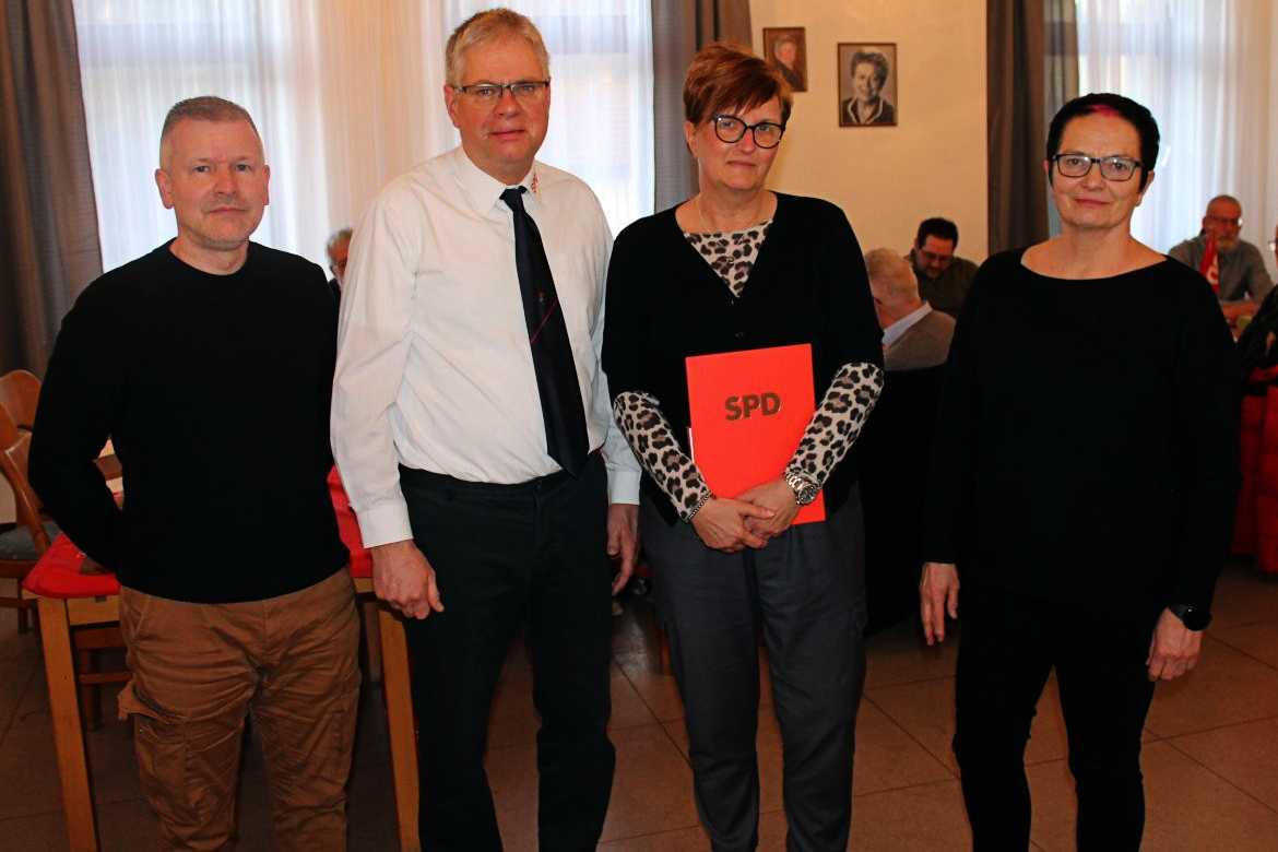 Vier Personen stehen nebeneinander, eine Frau hält einen roten Ordner mit dem SPD-Logo. Im Hintergrund sind weitere Personen und ein Porträt zu sehen.