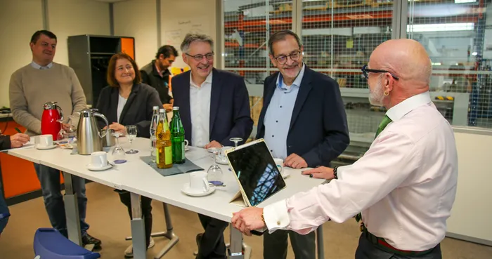 Eine Gruppe von fünf Personen steht um einen Tisch und diskutiert. Im Hintergrund ist ein Lager mit orangefarbenen Regalen zu sehen.