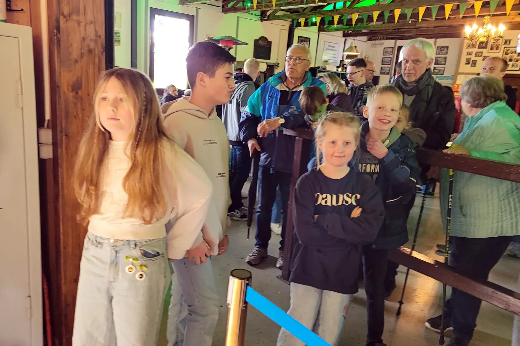 Mehrere Kinder und Erwachsene stehen in einem Raum mit Holztäfelung und blicken gespannt nach vorne, während ein Schützenfest eröffnet wird.