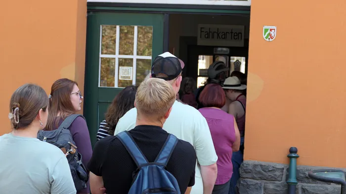 Menschen stehen vor einem Eingang mit der Aufschrift „Fahrkarten“ an einem orangefarbenen Gebäude.