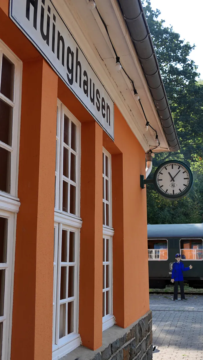 Orangefarbenes Gebäude mit dem Stationsschild „Hüinghausen“, einer Bahnhofsuhr und einem grünen Zug im Hintergrund.