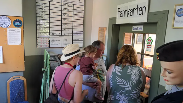Besucher stehen an einem Schalter mit der Aufschrift „Fahrkarten“ neben einem Fahrplan an der Wand.