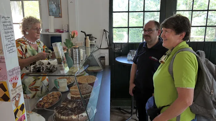Eine Verkäuferin hinter einer Glasvitrine mit verschiedenen Kuchen bedient zwei Kunden in einem Café.
