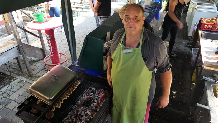 Ein Mann in einer grünen Schürze steht an einem Grill, auf dem Würstchen und Fleisch zubereitet werden.