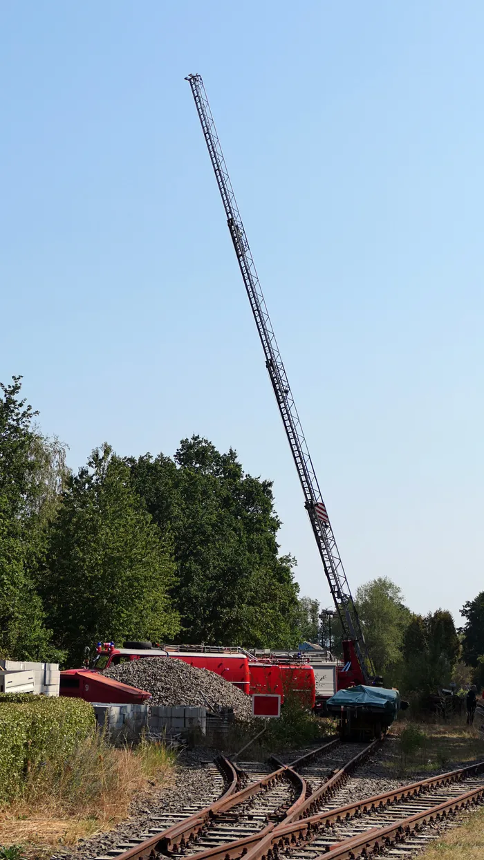 Ein rotes Schienenfahrzeug mit einem langen, hoch in den Himmel ragenden Gittermast auf Bahngleisen.