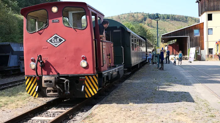 Eine rote O&K-Museumslokomotive mit grünen Waggons steht an einem Bahnsteig mit wartenden Besuchern.