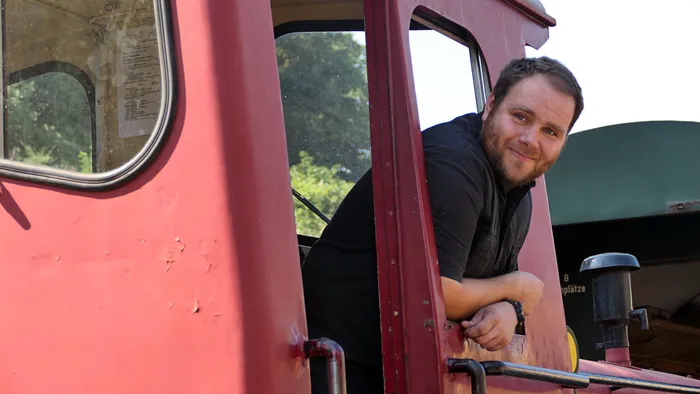 Ein bärtiger Mann in einem schwarzen Hemd lehnt lächelnd aus dem Fenster einer roten Lokomotive.