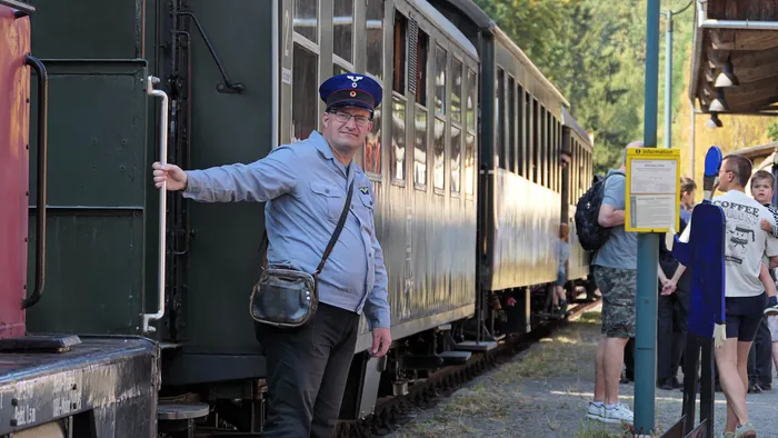 Ein Bahnmitarbeiter in blauer Uniform und Schirmmütze steht neben einem historischen Eisenbahnwaggon.