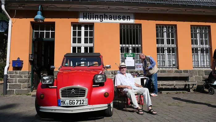 Ein roter Citroën 2CV steht vor einem orangefarbenen Gebäude mit der Aufschrift „Hüinghausen“, neben dem ein Mann auf einem Stuhl sitzt.