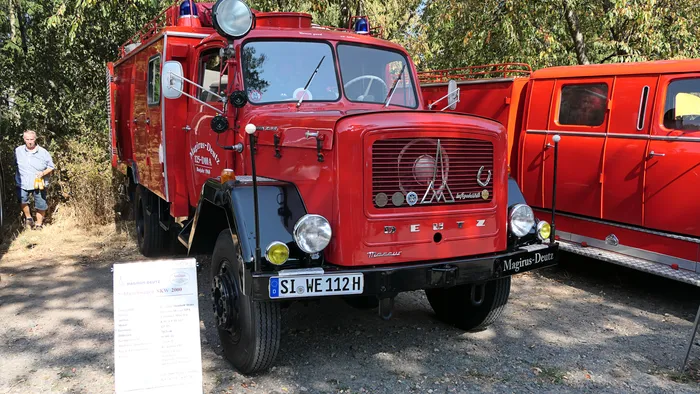 Ein rotes, historisches Magirus-Deutz Löschfahrzeug steht auf einem Schotterplatz vor Bäumen.