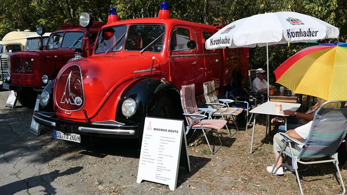 Ein rotes historisches Magirus-Deutz Löschfahrzeug steht neben Personen, die an Tischen unter Sonnenschirmen sitzen.
