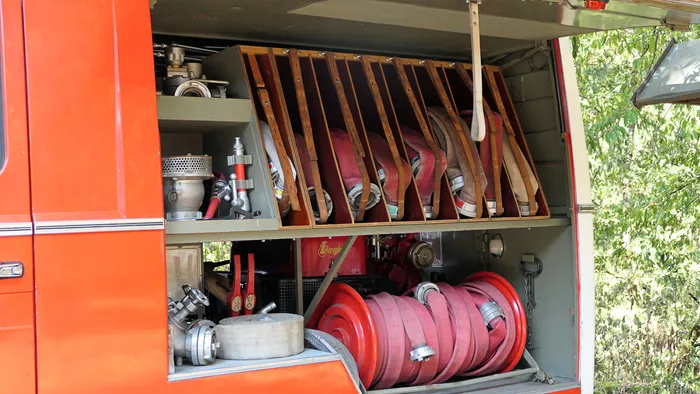 Offener Geräteraum eines roten Feuerwehrfahrzeugs mit Feuerwehrschläuchen in Holzregalen und einer großen roten Schlauchrolle.