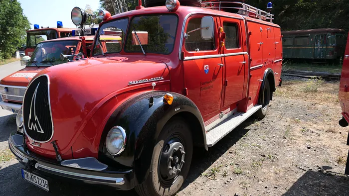 Ein rotes, historisches Magirus-Deutz Löschfahrzeug mit markantem silbernem Kühlergrill steht auf einem Schotterplatz.