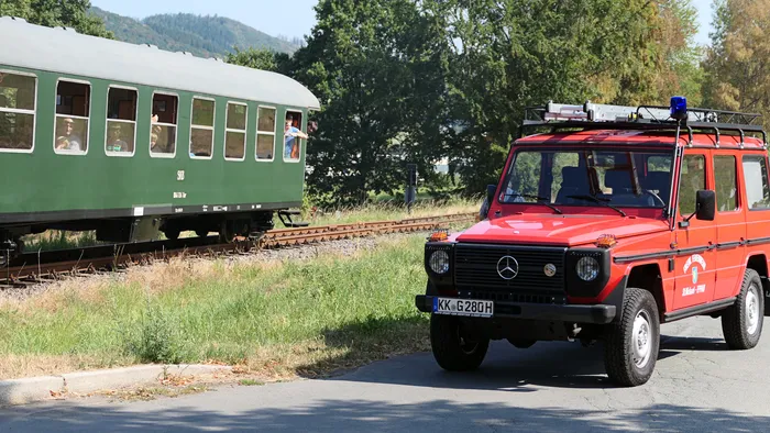 Ein rotes Feuerwehrfahrzeug fährt auf einer Straße vor einem grünen Museumszug auf Gleisen.