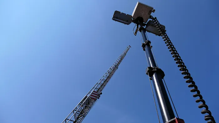 Ein schwarzer Lichtmast mit Scheinwerfern und einem Spiralkabel vor blauem Himmel, im Hintergrund eine Metallleiter.