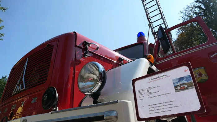 Nahaufnahme eines roten, historischen Magirus Deutz Feuerwehrfahrzeugs mit einem Informationsschild im Vordergrund.