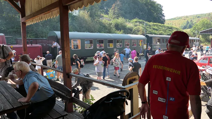 Besucher auf einem Bahnsteig neben einem historischen grünen Museumszug. Im Vordergrund steht ein Mann mit einem roten T-Shirt mit der Aufschrift „FEUERWEHR“.