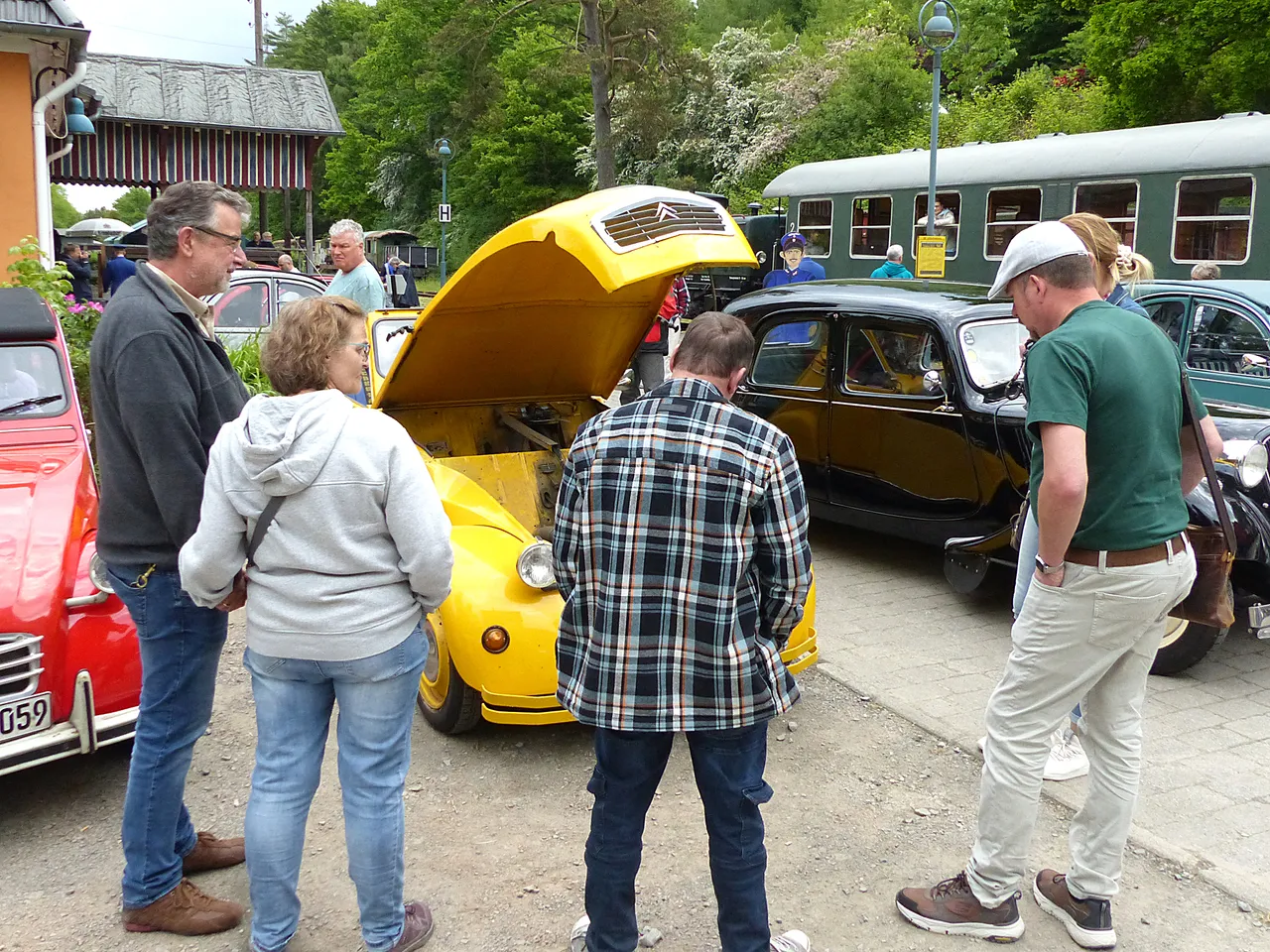 Mehrere Personen betrachten den geöffneten Motorraum eines gelben Citroën 2CV auf einem Platz vor historischen Eisenbahnwaggons.
