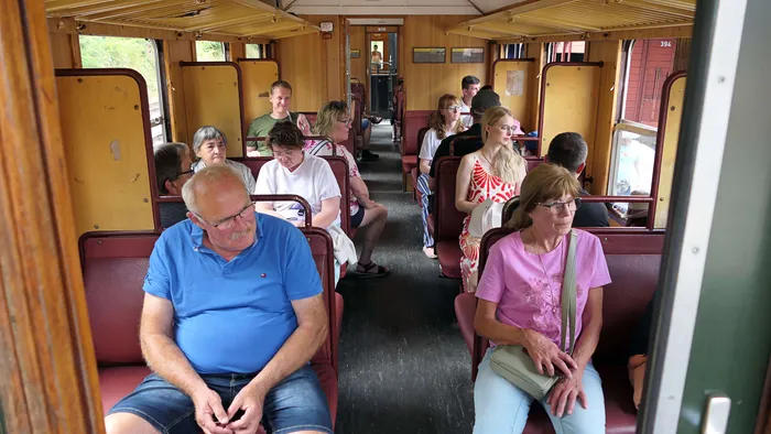 Passagiere sitzen und stehen in einem Waggon der Sauerländer Kleinbahn. Einige blicken aus den Fenstern, andere unterhalten sich oder sitzen entspannt.