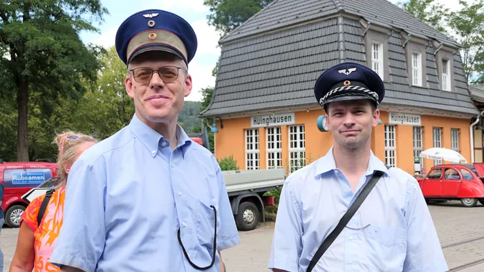 Zwei Männer in historischen Zugführeruniformen stehen vor dem Bahnhof Hüinghausen während eines Fahrtags der Sauerländer Kleinbahn.