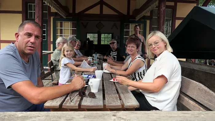 Eine Familie und weitere Personen sitzen an einem Holztisch vor einem Gebäude mit Fachwerk und genießen Getränke und Snacks während eines Fahrtags der Sauerländer Kleinbahn.