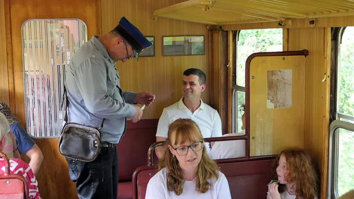 Ein Schaffner kontrolliert in einem Waggon der Sauerländer Kleinbahn die Fahrkarten einer Frau und eines Mannes, während im Hintergrund ein Kind schaut.