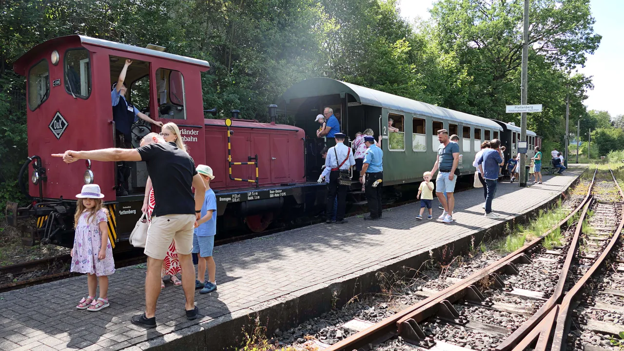 Eine rote Lokomotive der Sauerländer Kleinbahn steht auf Gleisen, umgeben von Fahrgästen. Einige Personen zeigen in Richtung der Lokomotive, während andere bereits in den Waggons Platz genommen haben.