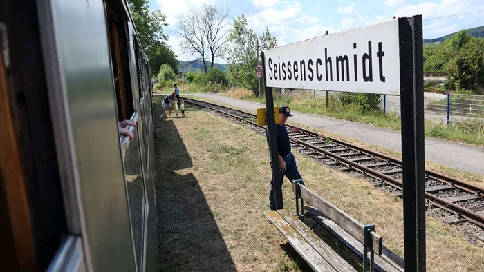 Ein Mitarbeiter der Sauerländer Kleinbahn steht neben einem Bahnschranken und einem Schild mit dem Ortsnamen Seisenschmidt, während Fahrgäste aus einem Zugfenster schauen.