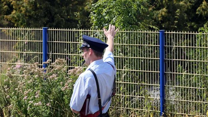 Ein Mitarbeiter der Sauerländer Kleinbahn steht mit erhobener Hand vor einem blauen Maschendrahtzaun und weist in die Ferne.