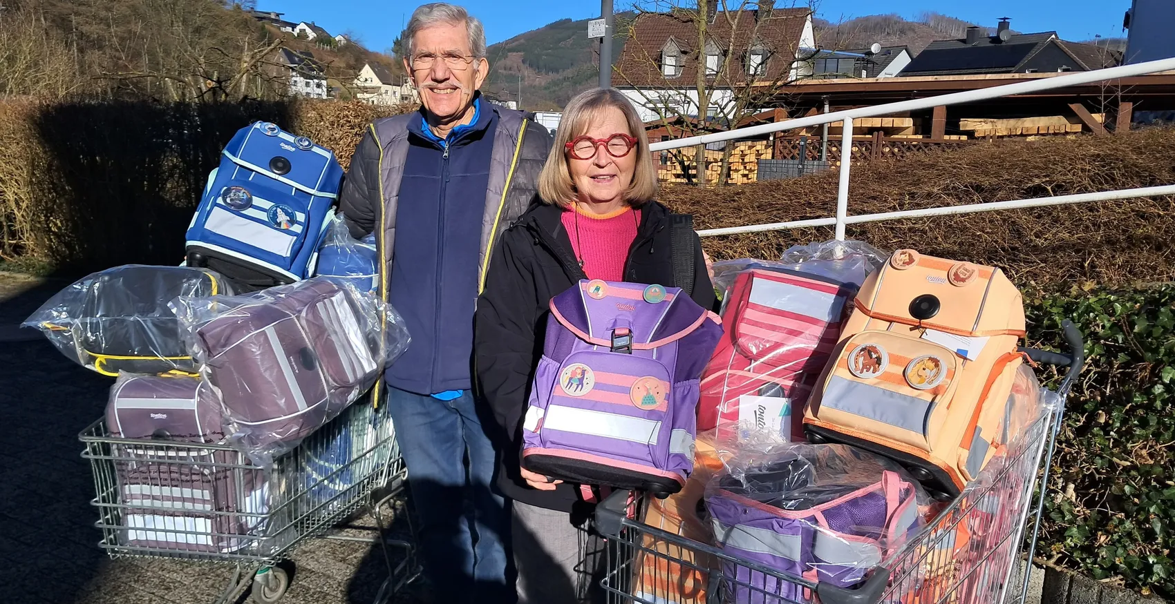 Ein älteres Ehepaar steht vor einem Einkaufswagen und einem Anhänger, beide voll mit neuen Schulranzen in verschiedenen Farben und Designs. Sie unterstützen das Projekt „Guter Schulstart“ für Kinder aus finanziell benachteiligten Familien.