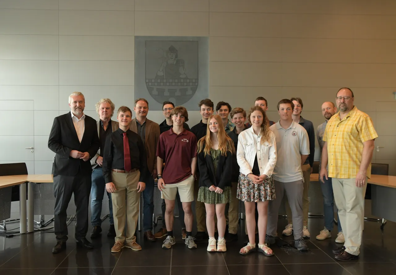 Eine Gruppe von Jugendlichen und erwachsenen Männern steht im Ratsaal von Lüdenscheid vor einer großen, hellen Wand mit einem Wappen. Bürgermeister Sebastian Wagemeyer steht links im Bild in einem Anzug und begrüßt die Schüler aus Fort Wayne.