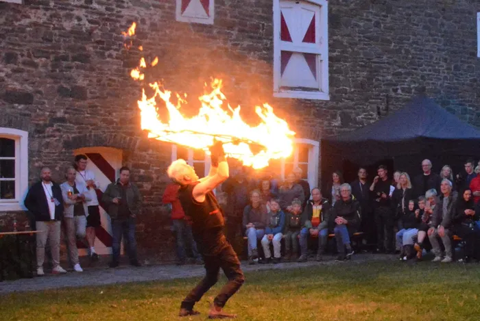 Ein Mann führt eine Feuershow vor einem historischen Gebäude mit Zuschauern auf einer Rasenfläche auf.