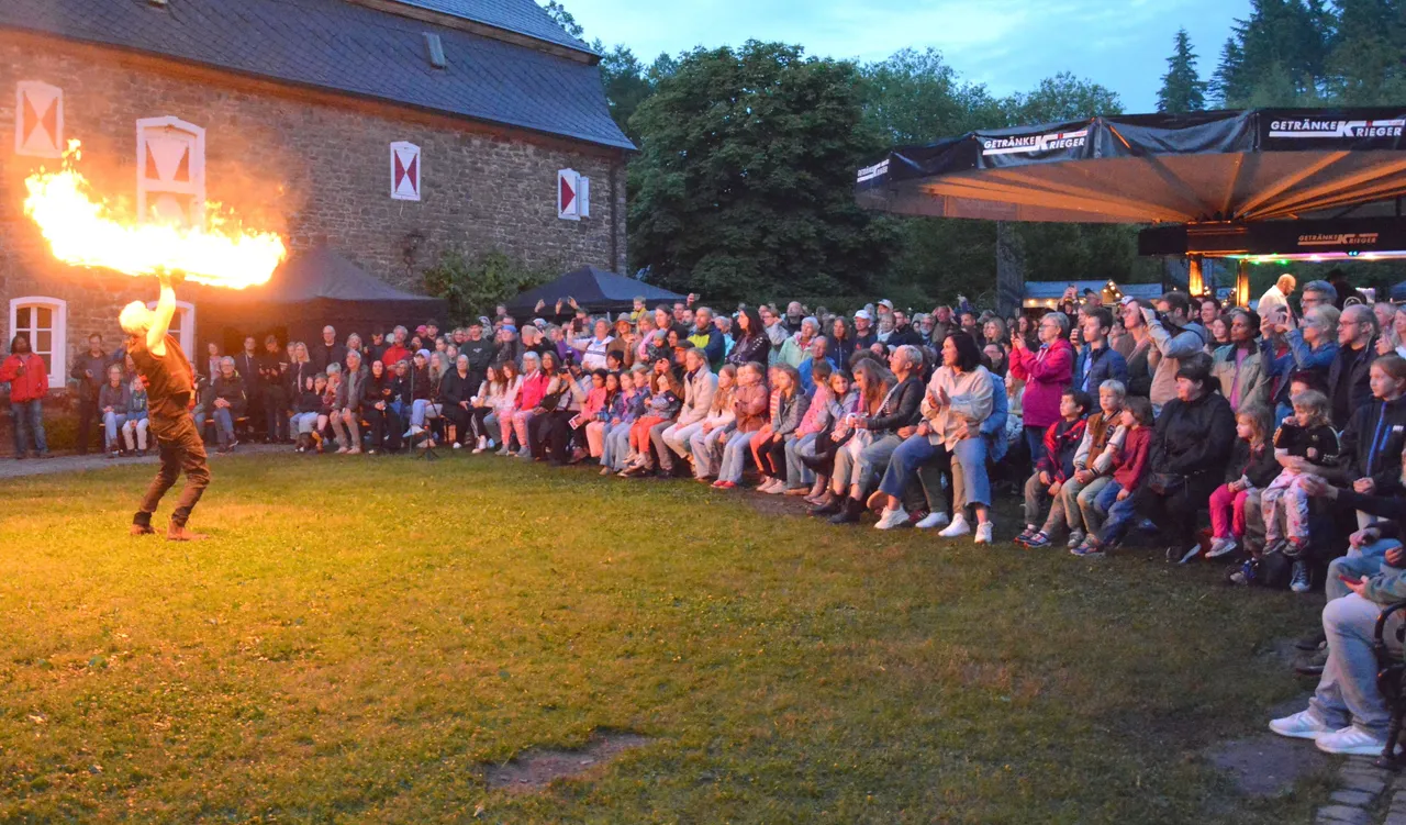 Feuershow vor großem Publikum auf dem Gelände von Schloss Neuenhof. Ein Künstler jongliert mit brennenden Keulen, während Zuschauer auf Bänken und Stühlen sitzen.